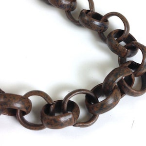 Puede incluir: Primer plano de una sección de una cadena grande de color marrón oxidado. Los eslabones ovalados tienen un aspecto texturizado y desgastado. La cadena se encuentra sobre un fondo blanco brillante, resaltando su color y textura.