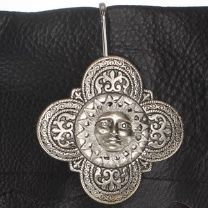 Può includere: Uncino decorativo in metallo argentato a forma di sole con dettagli decorativi. L'uncino è attaccato a una superficie in pelle nera.