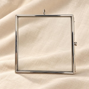 May include: A square, silver-toned metal frame with clear glass panels. The frame features a thin, polished metallic border, a small circular hanging loop at the top centre, and a side-mounted latch mechanism for opening and closing the display.
