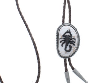 Può includere: Un cravattino bolo in pelle marrone con un pendente argentato che presenta uno scorpione nero su sfondo bianco.