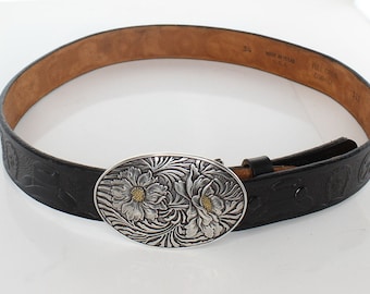 May include: Black leather belt with a silver and gold floral buckle. The buckle is oval shaped and has a detailed design. The belt is embossed with a floral pattern. The belt is labelled "Full Grain Cowhide" and "Made in Texas".