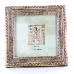 May include: Square framed artwork featuring a pink ribbon made of sparkling gems. The artwork is set against a light green background with the words "Let Your Light Shine" below the ribbon. The frame is a light brown color.