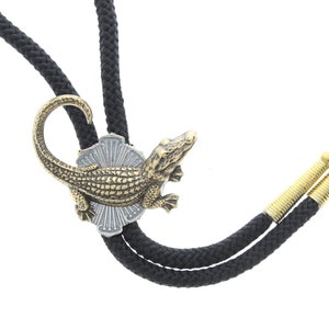 May include: A black bolo tie with gold-toned tips and a decorative alligator pendant. The alligator is detailed with scales and has a curled tail.