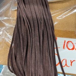 May include: A dark brown suede tassel with numerous thin, fringed strands. The tassel is a rich, earthy colour and appears to be made of soft material. The tassel is likely used for decorative purposes, such as on a handbag or keychain.