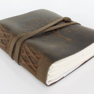 May include: A brown leather journal with a tree design embossed on the cover. The journal has a leather strap closure and is tied with a braided leather cord.