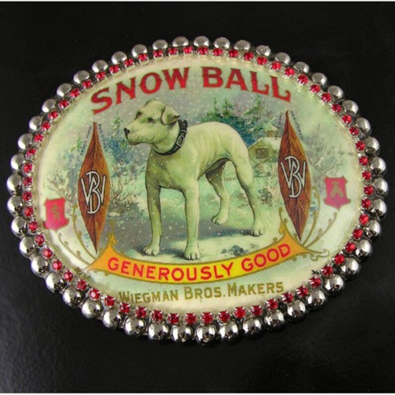 May include: A silver and red oval belt buckle with a white dog on a snowy background. The buckle features the text "SNOW BALL GENEROSELY GOOD WIEGMAN BROS. MAKERS".