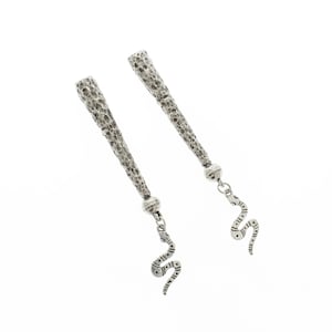 Può includere: Un paio di orecchini argentati. Ogni orecchino presenta una parte superiore cilindrica strutturata, una piccola sfera e un ciondolo a forma di serpente pendente. Il ciondolo a serpente ha un design a motivi. Gli orecchini sono su sfondo bianco.