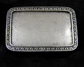 May include: A silver-toned metal belt buckle with a rectangular shape and a decorative border. The buckle has a smooth, blank surface.