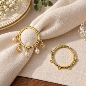 May include: Two circular gold-toned napkin rings with textured edges. One ring features four dangling charms, including two white spherical pearls and two faceted clear crystal beads. The second ring has four empty loops for attaching charms.