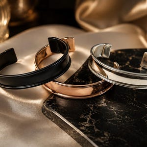 May include: Three open cuff bracelets featuring a smooth, curved design. One bracelet is matte black, one is polished rose gold, and one is polished silver. Each bracelet has a wide, slightly concave center band with raised edges.