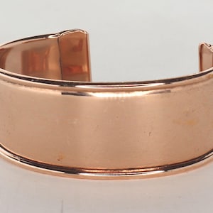 May include: A wide copper cuff bracelet with a smooth, polished finish. The bracelet has a rounded edge and a slightly curved shape.