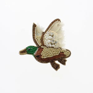 May include: A beaded duck brooch in flight, featuring a green head, brown and tan body, and white accents on the wings. The brooch is embellished with beads and sequins, creating a textured and detailed design.