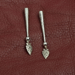 Puede incluir: Un par de pendientes de plata con un diseño de punta de flecha martillada. Los pendientes son largos y delgados, con una pequeña perla de plata en la parte superior.