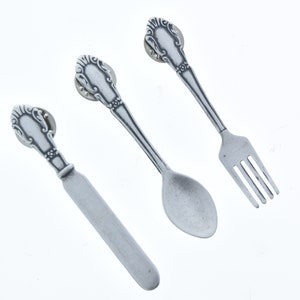 May include: A set of three silver-toned metal miniature utensils: a fork, a spoon, and a butter knife. Each utensil has an ornate, decorative handle.