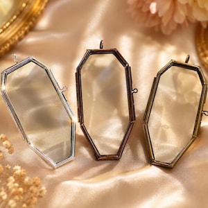 Puede incluir: Tres marcos de cristal con forma de ataúd y bordes metálicos en acabados plateado, cobre y latón. Cada marco cuenta con un cierre de bisagra, una pequeña anilla superior para colgar y paneles de cristal transparente en un borde metálico.