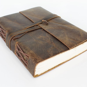 May include: A brown leather journal with a leather strap closure and decorative stitching. The journal is closed and lying on a white surface.