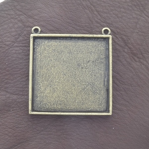 May include: A square-shaped metal pendant with a gold finish. The pendant has two loops at the top for attaching a chain or cord. The pendant is designed to hold a square-shaped piece of glass or other material.