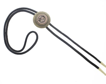 May include: A bolo tie with a circular, bronze-colored pendant featuring a detailed design. The pendant is attached to a black cord with gold-colored tips. The tie is set against a white background.