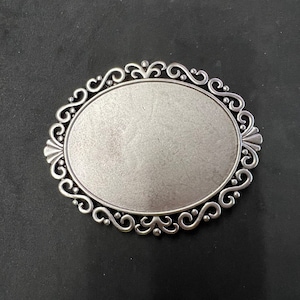 Puede incluir: Un marco de metal plateado de forma ovalada con un diseño decorativo de desplazamiento. El marco está en blanco y listo para ser personalizado.