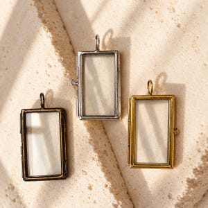 May include: Three rectangular glass pendants with metal frames in bronze, silver, and gold tones. Each pendant features a hinged design with a small loop at the top for hanging and a side latch, providing a clear display space for small items.