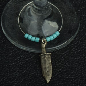 Peut inclure: Un porte-verre à vin avec un pendentif en forme de couteau argenté. Le charme est orné de perles turquoise et est suspendu à un anneau en fil d'argent. Le pendentif en forme de couteau a une surface texturée et une poignée détaillée.