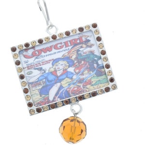 Peut inclure: Un charme argenté avec une couverture de bande dessinée vintage représentant une femme en chemise bleue et un chapeau de cow-boy. Le charme est entouré de strass marrons et a un cristal ambré suspendu.