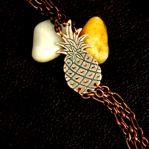 May include: A copper pineapple charm hangs from a multi-strand copper chain. The charm is detailed with a textured pineapple design. The chain is attached to two small, smooth stones.