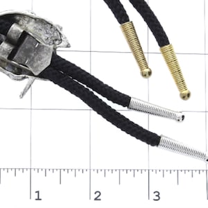 May include: A silver bolo tie with a detailed design of a Native American headdress. The tie has black cords with silver and gold tips.