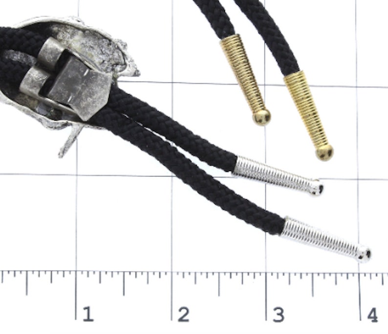 May include: A silver bolo tie with a detailed design of a Native American headdress. The tie has black cords with silver and gold tips.