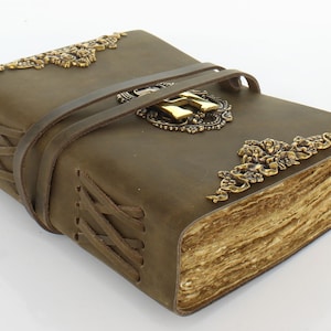 May include: An olive-green leather journal with aged paper pages. It has brass corner accents and a central emblem. A leather strap secures the journal. The edges are stitched with a crisscross pattern. The journal is approximately 20 cm in length.