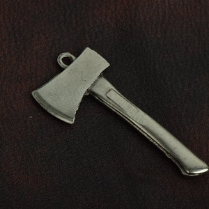 Puede incluir: Colgante de hacha plateada con un lazo para colgar. La cabeza del hacha tiene una hoja ligeramente curvada, y el mango presenta un detalle rectangular. Sobre un fondo marrón oscuro con aspecto de cuero.