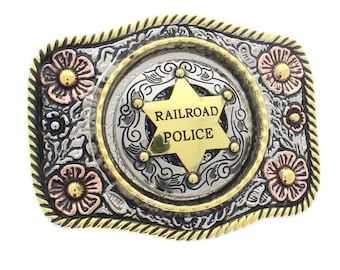 Puede incluir: Una hebilla de cinturón de color dorado, plateado y cobre con un diseño ornamentado. La hebilla presenta una estrella de cinco puntas con las palabras "Railroad Police" en su interior. La hebilla está rodeada de diseños florales.