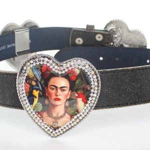May include: Black leather belt with a silver buckle featuring a heart-shaped design with a portrait of Frida Kahlo surrounded by birds and flowers. The belt has three heart-shaped buckles.