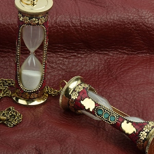 May include: Two ornate gold-toned hourglass pendants with red and green accents. The hourglasses are on a red leather surface. One hourglass has a chain attached.