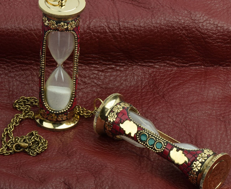 May include: Two ornate gold-toned hourglass pendants with red and green accents. The hourglasses are on a red leather surface. One hourglass has a chain attached.