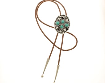 May include: A bolo tie with a brown leather cord and a silver-toned oval pendant. The pendant features five turquoise-colored stones and decorative silver filigree. The tie has silver-toned tips.