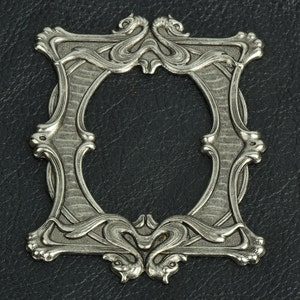 May include: A silver-toned metal frame featuring an ornate, symmetrical design with stylized dragon or serpent motifs at the top and bottom. The central opening is oval-shaped, surrounded by textured, grooved detailing and curved, flowing lines.