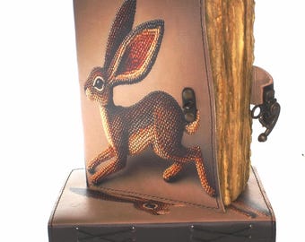 Pode incluir: Um diário de couro marrom com um desenho de lebre correndo. O diário tem um fecho decorativo e páginas envelhecidas. O diário está em cima de uma caixa de couro combinando com costuras decorativas.