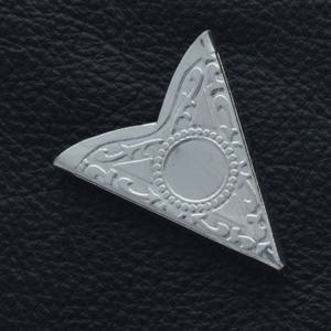 Puede incluir: Protección de esquina de metal plateada con un diseño decorativo. El protector de esquina tiene una forma triangular con un centro circular.