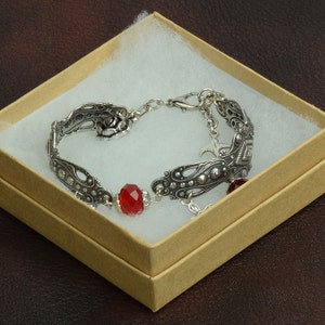 Pode incluir: Um bracelete de prata com um design de rosa, contas vermelhas e um amuleto de pássaro. O bracelete está em uma caixa marrom com acolchoamento branco.
