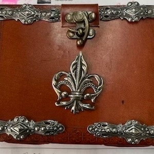 Puede incluir: Un diario encuadernado en cuero marrón con adornos de metal plateado ornamentados, incluyendo un diseño de flor de lis y un cierre de broche.
