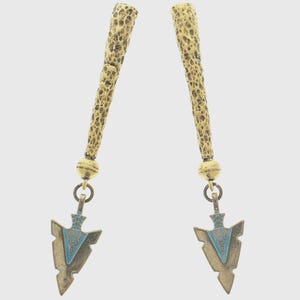 Puede incluir: Un par de pendientes dorados. Cada pendiente presenta una parte superior cilíndrica texturizada, una pequeña cuenta dorada y un colgante en forma de punta de flecha con detalles de esmalte azul. Fondo blanco.