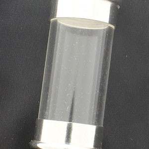 Puede incluir: Un colgante de plata y vidrio transparente con un bucle en la parte superior. El colgante tiene forma cilíndrica y un acabado liso y pulido.