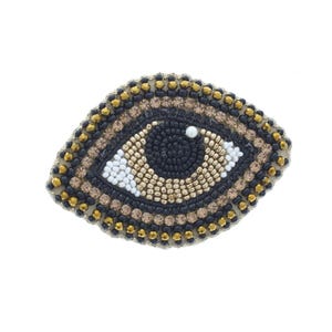 May include: An eye-shaped patch featuring intricate beadwork in black, gold, and white. The central pupil is composed of dark beads with a single white accent, surrounded by concentric layers of metallic gold, tan, and black beads in a textured pattern.