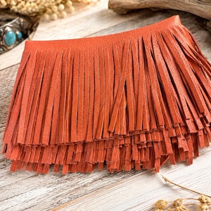 May include: A vibrant orange leather accessory featuring multiple layers of long, thin, vertical fringe strips. The material has a distinct pebbled texture, and the design consists of a solid, rectangular top section that transitions into dense, flowing fringe.