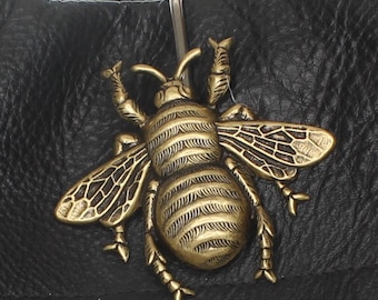 Può includere: Primo piano di un articolo in pelle nera con un emblema decorativo a forma di ape color oro antico. L'ape ha ali e segmenti del corpo dettagliati ed è visibile una chiusura in metallo color argento.