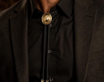 Puede incluir: Una corbata de bolo con una pieza central circular de metal dorado con un acabado liso y reflectante. La corbata consiste en un cordón trenzado negro que cuelga, terminado con dos herretes de metal dorado a juego en los extremos inferiores.