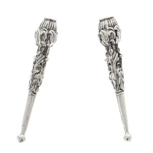 May include: A pair of silver-toned decorative finials with intricate floral designs. The finials are shaped like a pointed cone with a rounded top.