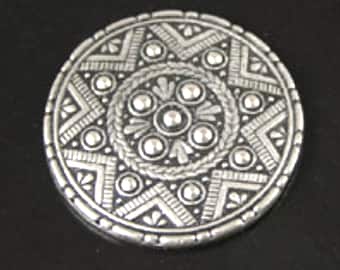 Op de afbeelding: Een zilveren ronde broche met een reliëf, ingewikkeld ontwerp. Het midden heeft een cirkelvormig patroon met een reliëf, getextureerde rand. De buitenrand van de broche is ook getextureerd.