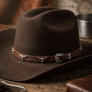 Op de afbeelding: Een donkerbruine vilten cowboyhoed met een bruine leren hoedband. De band is versierd met zilverkleurige metalen accenten in de vorm van stierenschedels en een zilverkleurige metalen gespsluiting. De hoed heeft een ingedeukte kroon en een brede, platte rand.